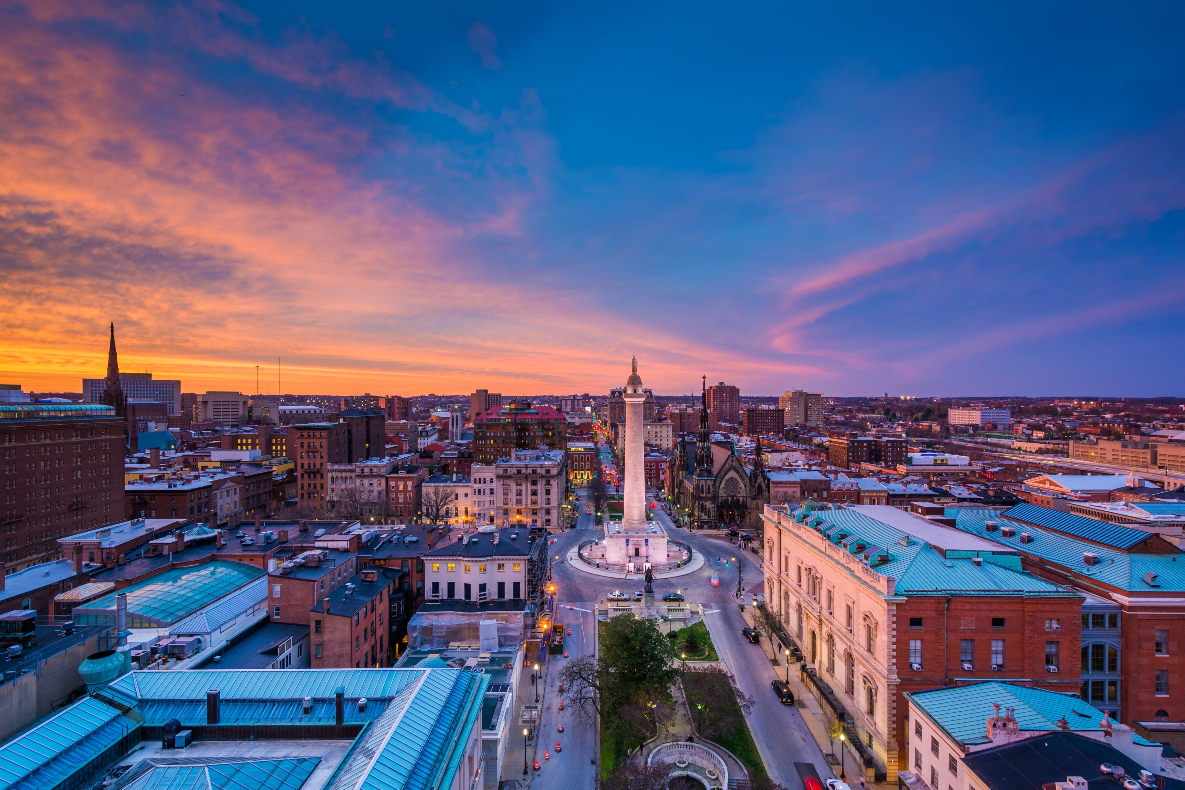 A cityscape at sunset with a prominent monument in the center, surrounded by historic buildings and a clear sky.