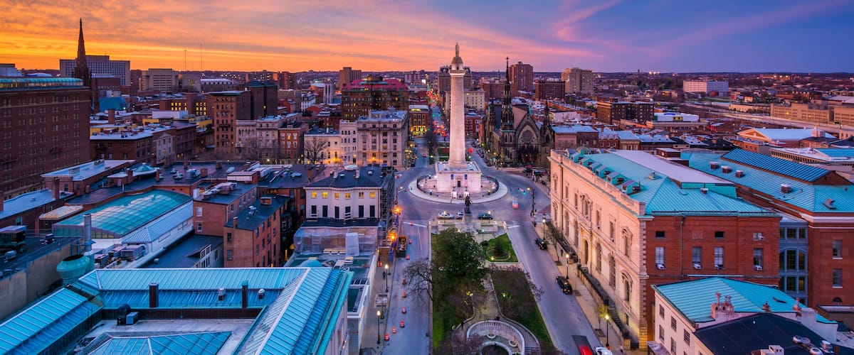 A cityscape at sunset with a prominent monument in the center, surrounded by historic buildings and a clear sky.