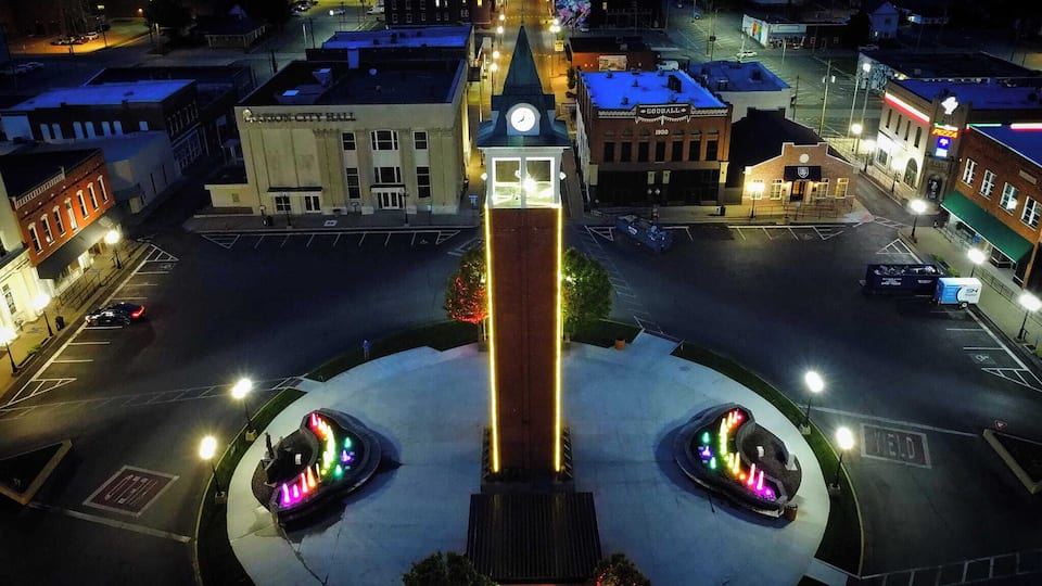 A city square with a central monument, surrounding buildings, and a parking lot.