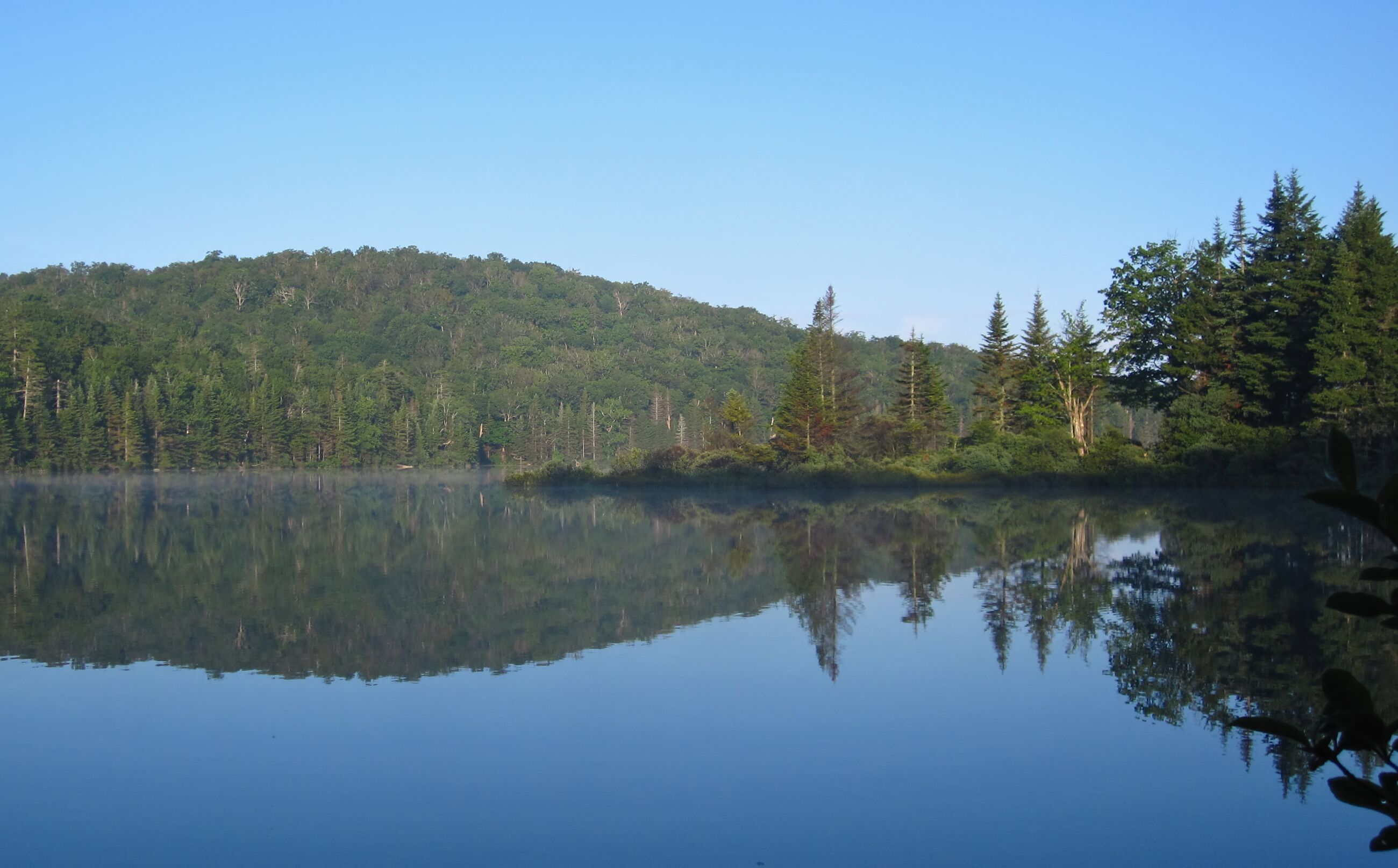 A calm lake with reflections of trees and a forested hill.