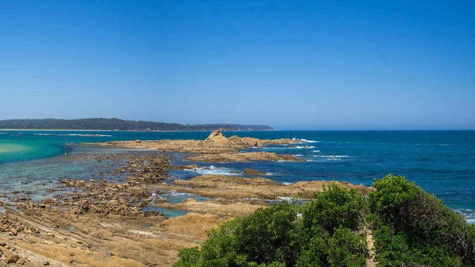 A coastal landscape with a sandy beach, turquoise waters, rocky outcrops, and green hills in the background.