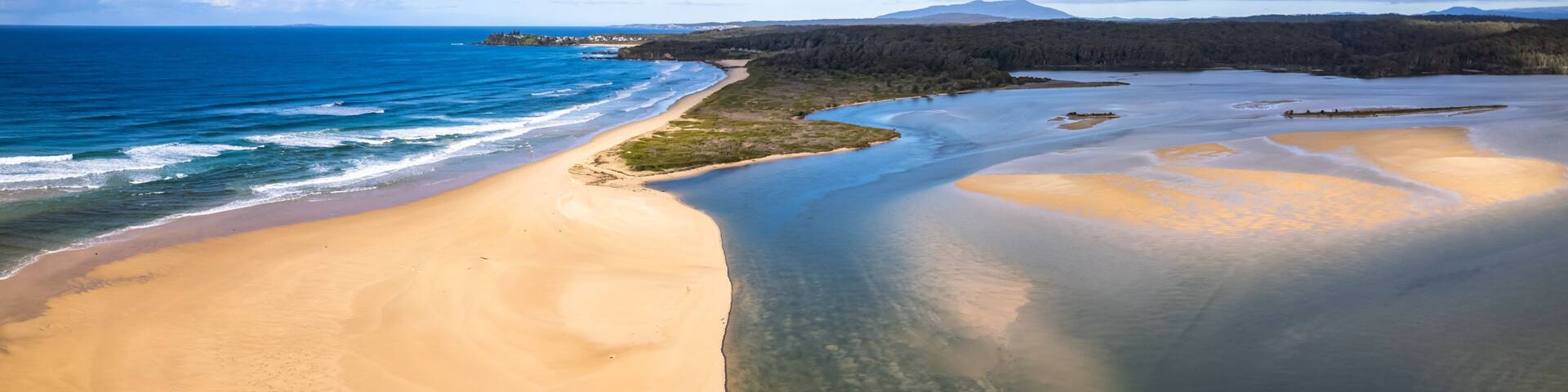 A coastal landscape with a sandy beach, clear blue waters, and a forested area in the background.