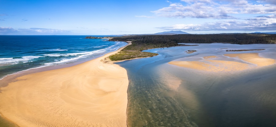 A coastal landscape with a sandy beach, clear blue waters, and a forested area in the background.