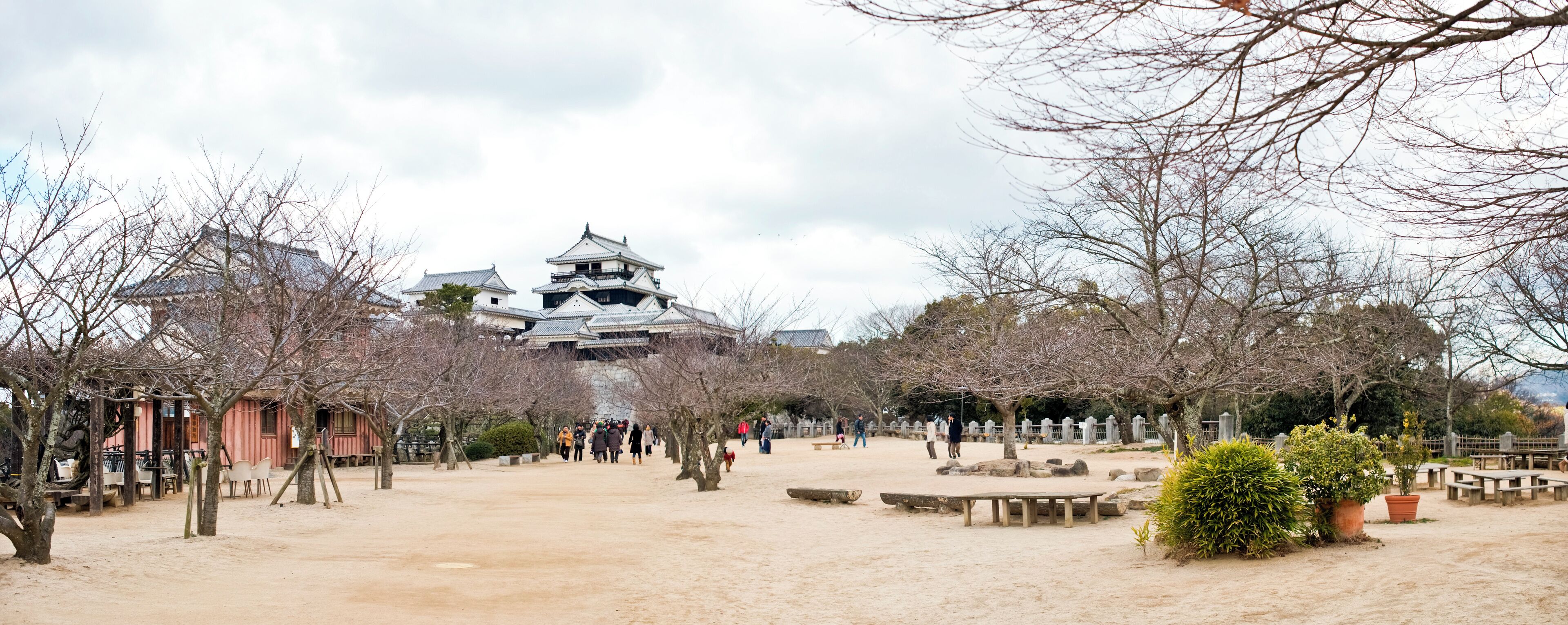A traditional Japanese castle with multiple tiers and a large central tower, surrounded by a sandy courtyard with trees and benches.