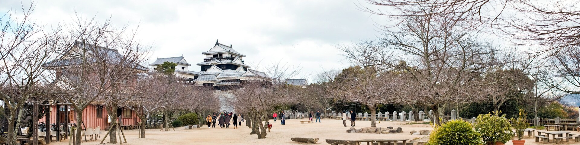 A traditional Japanese castle with multiple tiers and a large central tower, surrounded by a sandy courtyard with trees and benches.