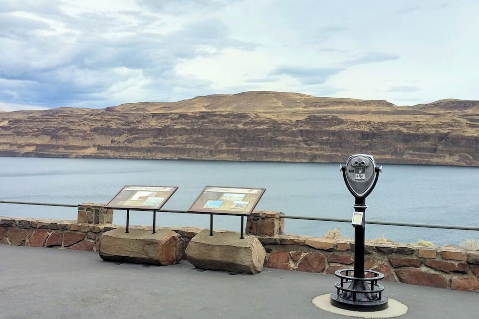 A scenic overlook with a telescope, two informational signs, and a lake with hills in the background.