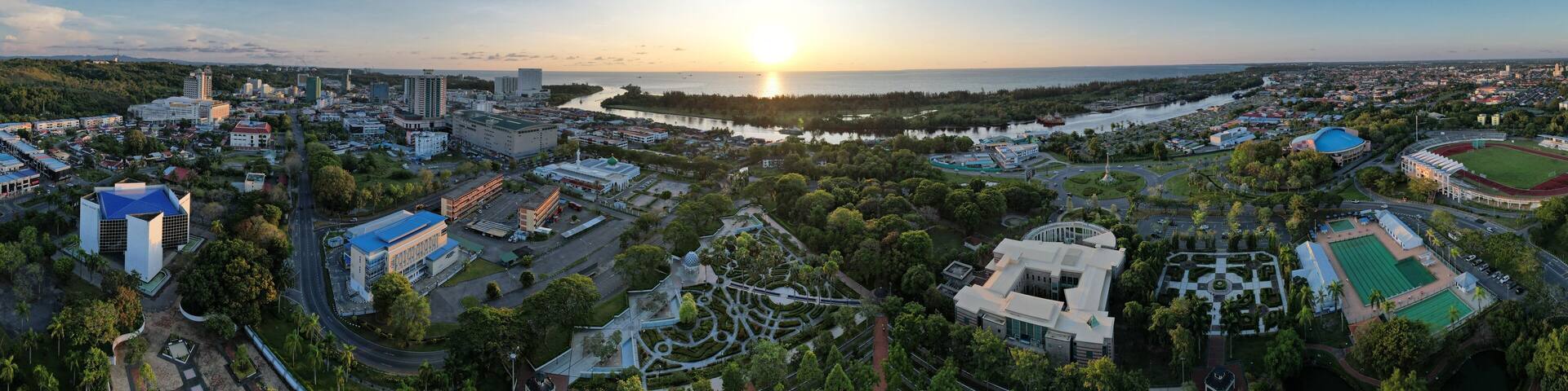A coastal cityscape at sunset with a river, buildings, and a park.