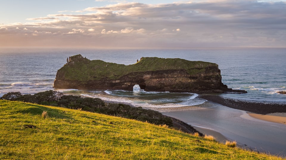 A coastal landscape with a prominent rock formation, grassy hills, and a beach.