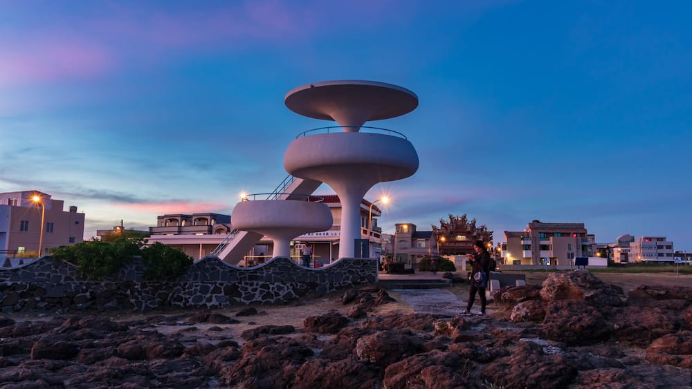 A modern architectural structure with a circular design, surrounded by rocky terrain and residential buildings in the background.