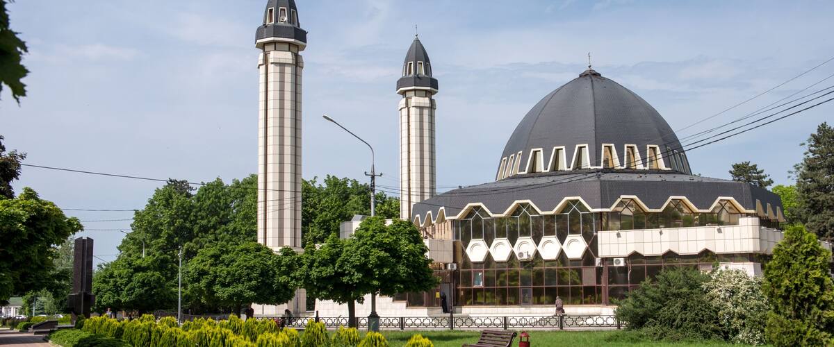 A mosque with a large dome and two minarets, surrounded by a landscaped garden with benches and a paved walkway.