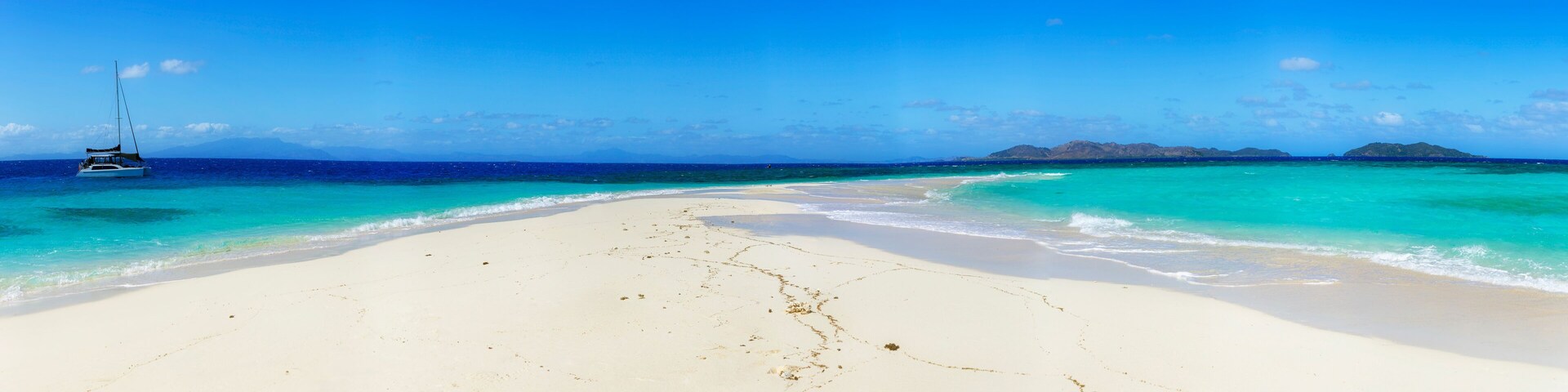 Praia de areia branca com águas turquesa, um veleiro e ilhas ao longe.