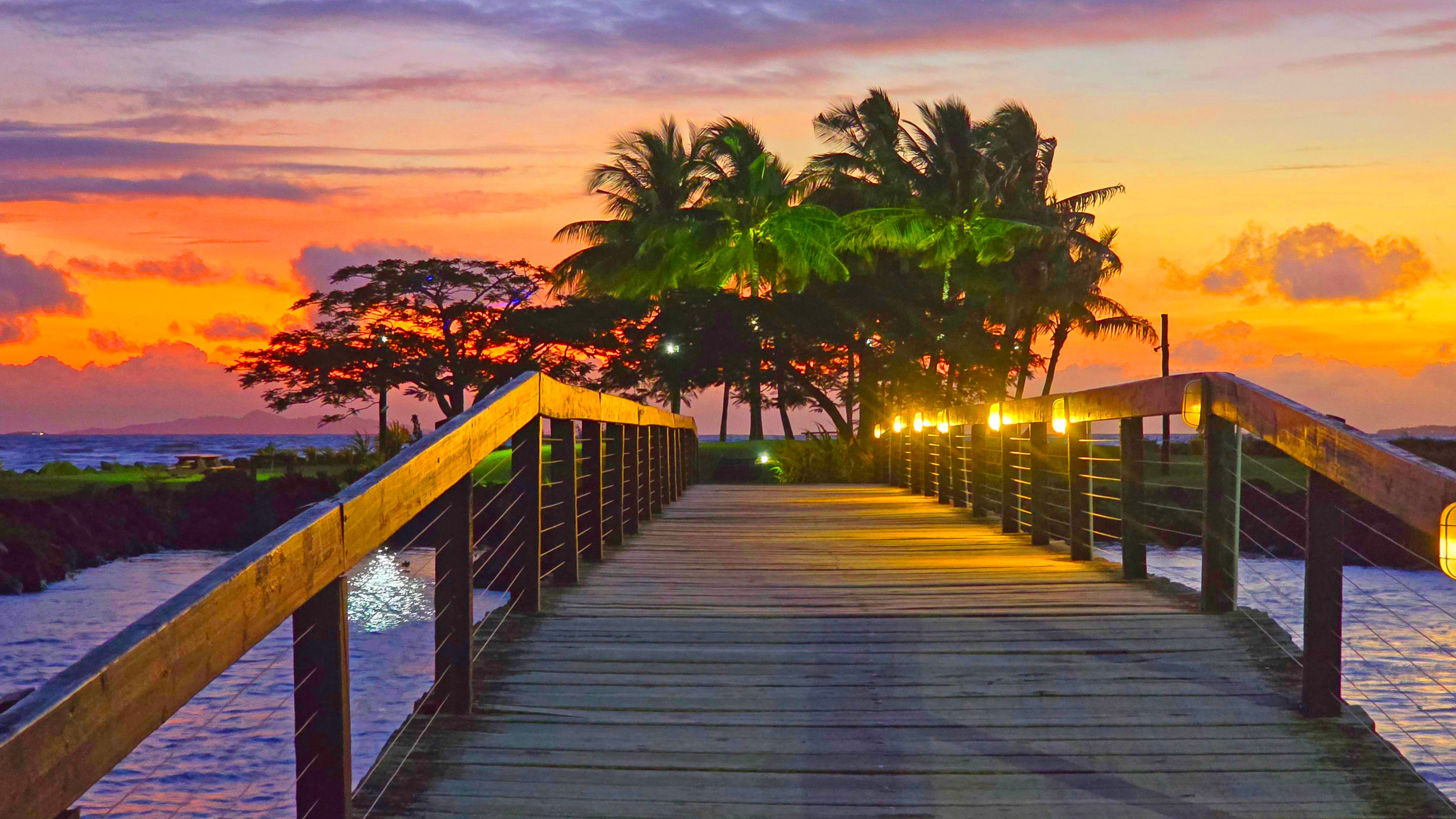 Una pasarela de madera que conduce a una playa con palmeras y un atardecer.