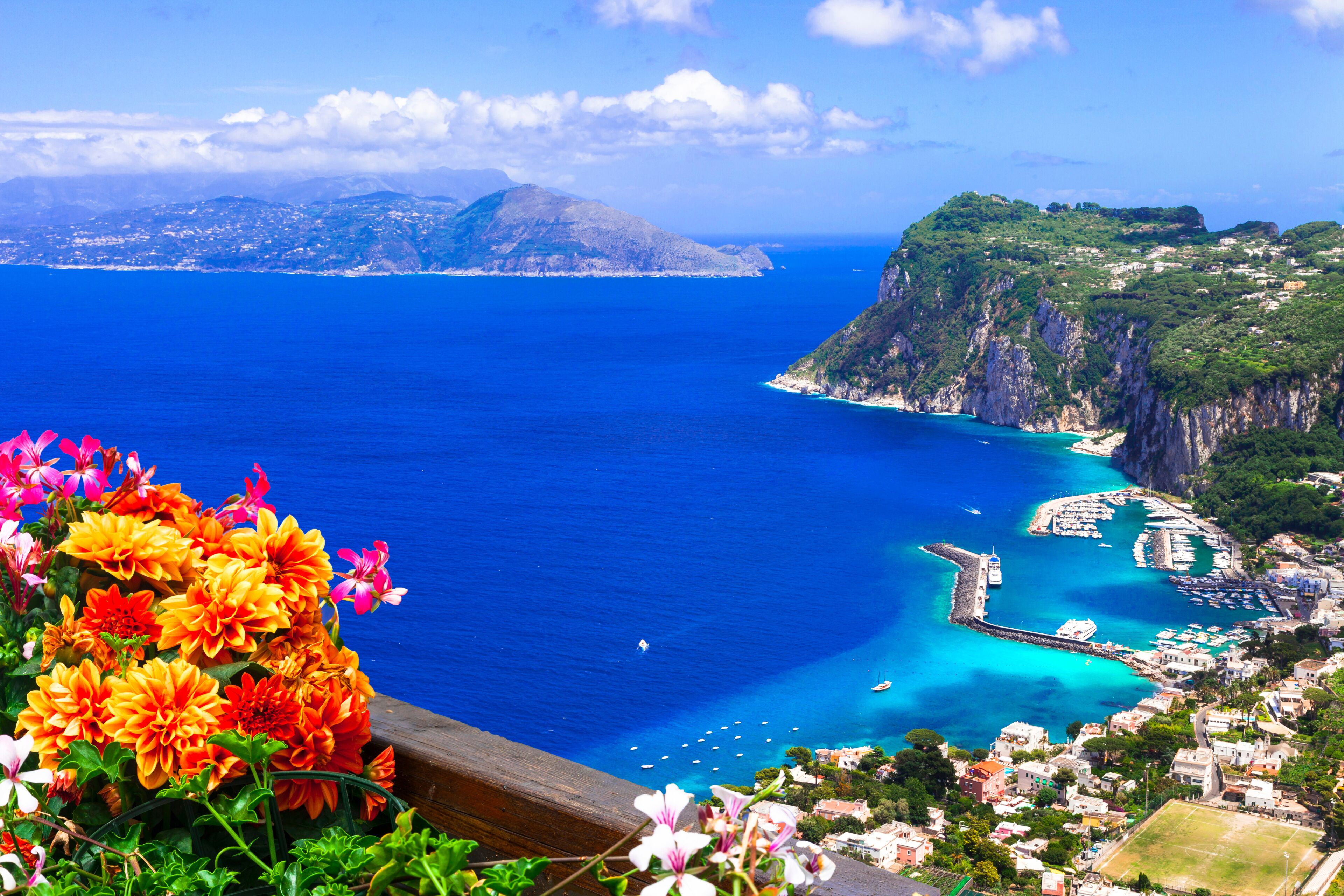 Paisagem costeira com mar azul-turquesa, um penhasco rochoso e um pequeno porto com barcos.