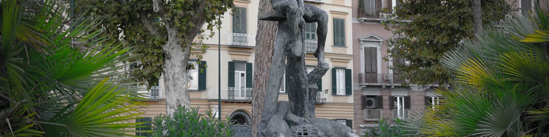 Estátua de uma figura sentada sobre base de tronco de árvore, cercada por palmeiras e edifícios ao fundo.
