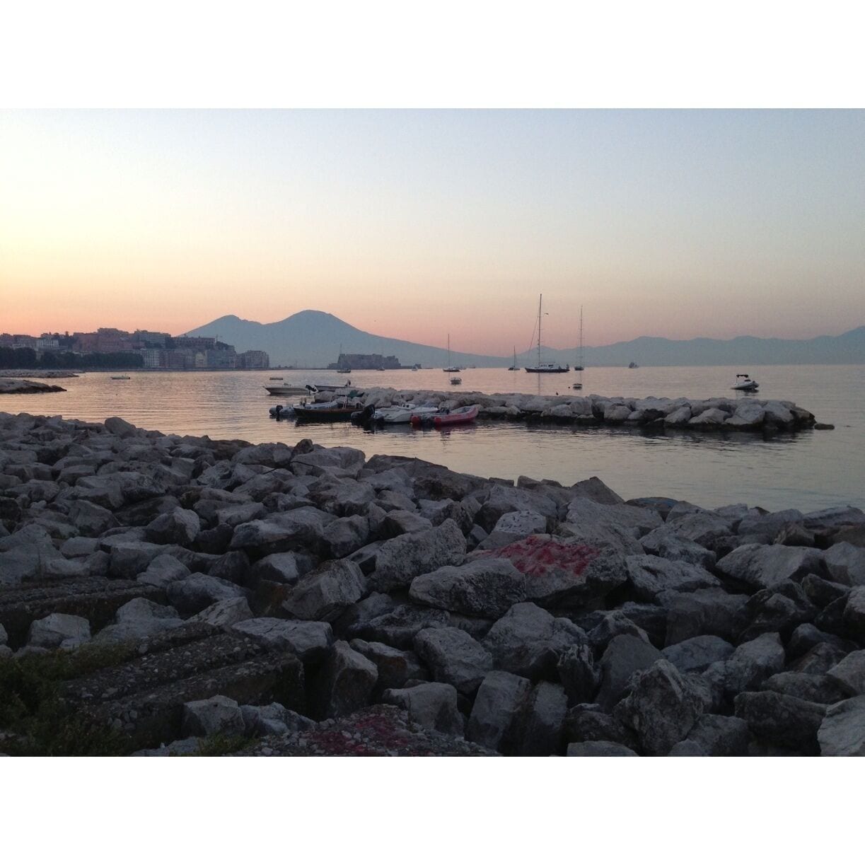 A rocky shoreline with boats docked at a pier, a distant mountain range, and a calm sea at sunset.