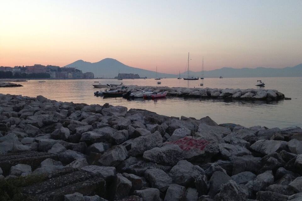 Costa rocosa con barcos amarrados en un muelle, una cadena montañosa distante y un mar tranquilo al atardecer.
