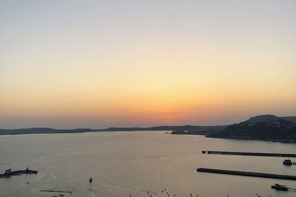 Un paysage côtier au coucher du soleil, avec une vaste étendue d’eau, une jetée et une colline au loin.