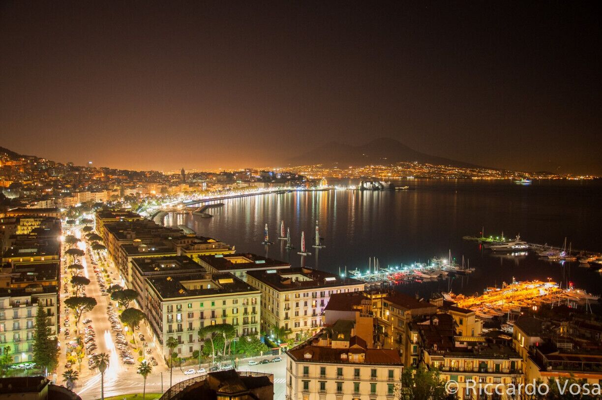 The beauty of Naples during the America's Cup World Series, in 2013. 
In front you can see Castel Dell'Ovo and beyond the silhouette of the active volcano Vesuvio. 

Naples' historic city centre is the largest in Europe, covering 1,700 hectares (4,200 acres) and enclosing 27 centuries of history, and is listed by UNESCO as a World Heritage Site. Naples has long been a major cultural centre with a global sphere of influence, particularly during the Renaissance and Enlightenment eras. In the immediate vicinity of Naples are numerous culturally and historically significant sites, including the Palace of Caserta and the Roman ruins of Pompeii and Herculaneum. Culinarily, Naples is synonymous with pizza, which originated in the city. Neapolitan music has furthermore been highly influential, credited with the invention of the romantic guitar and the mandolin, as well as notable contributions to opera and folk standards. Popular characters and historical figures who have come to symbolise the city include Januarius, the patron saint of Naples, the comic figure Pulcinella, and the Sirens from the Greek epic poem the Odyssey.