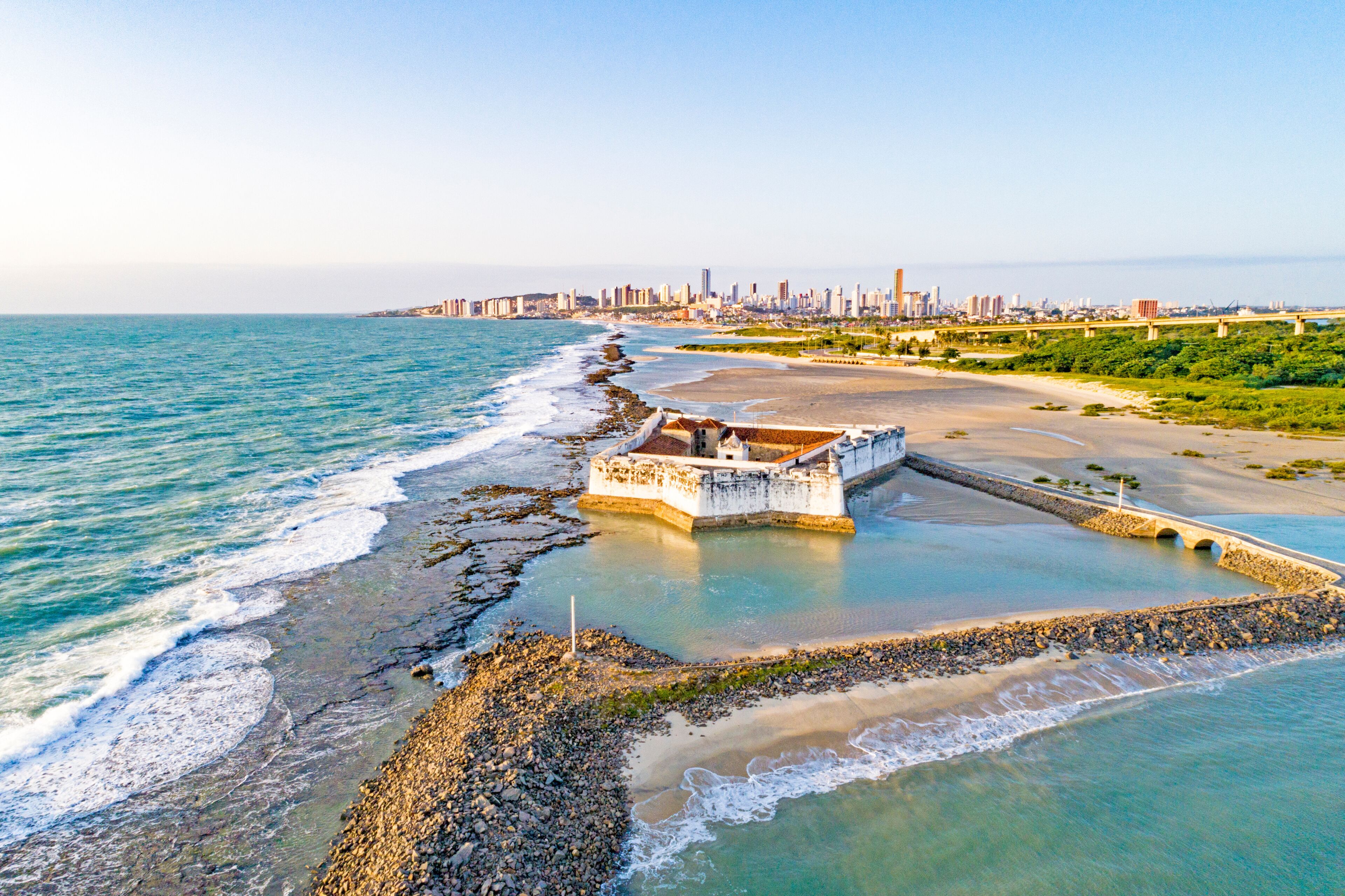 Un paisaje urbano costero con un muro marítimo destacado, una playa y un horizonte urbano lejano.