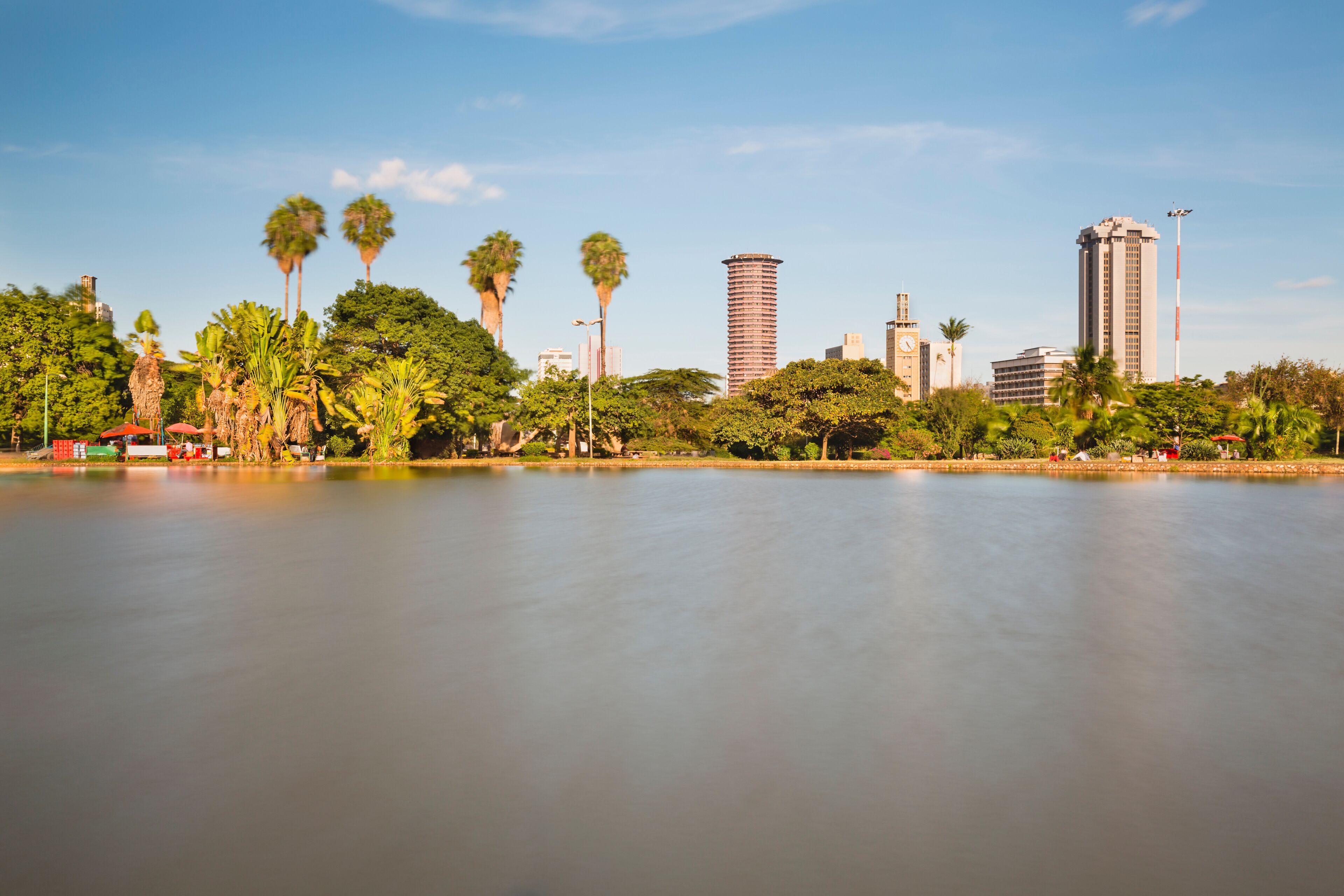 Un lago tranquilo con un horizonte urbano al fondo, que incluye palmeras y edificios modernos.