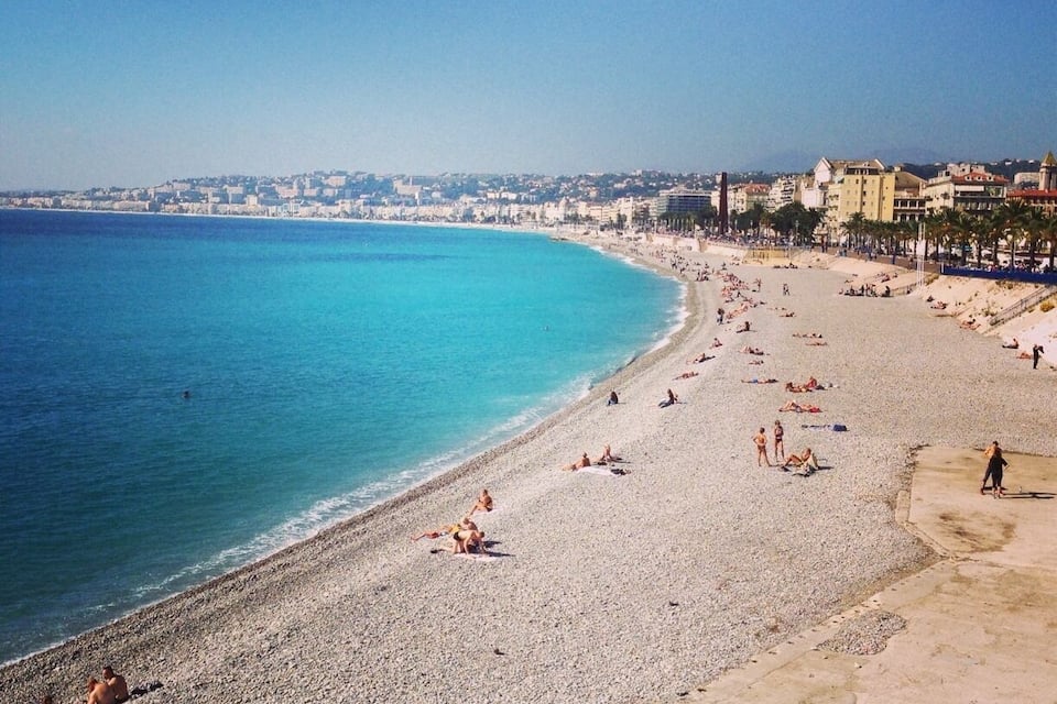 A pebble beach with people sunbathing and swimming, a clear blue sea, and buildings in the background.