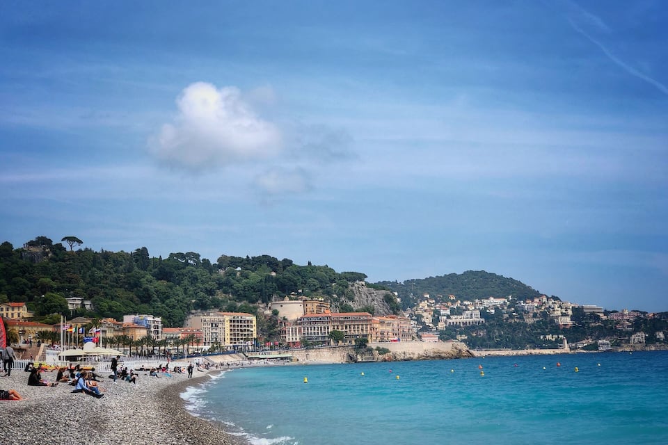 A pebble beach with people relaxing, a clear blue sea, and buildings on a hillside in the background.