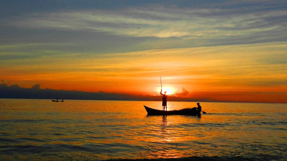 A sunset over a calm sea with two boats, one with a person standing and another with a person sitting.