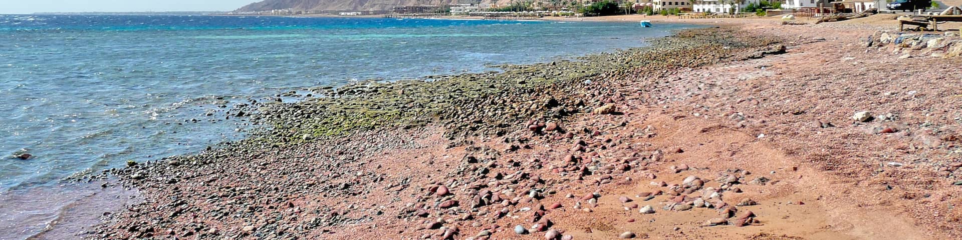 Coastal landscape north of Dahab, Sinai, Egypt, looking south — sunlit shoreline with distant buildings and the rugged mountain range rising beyond the Red Sea