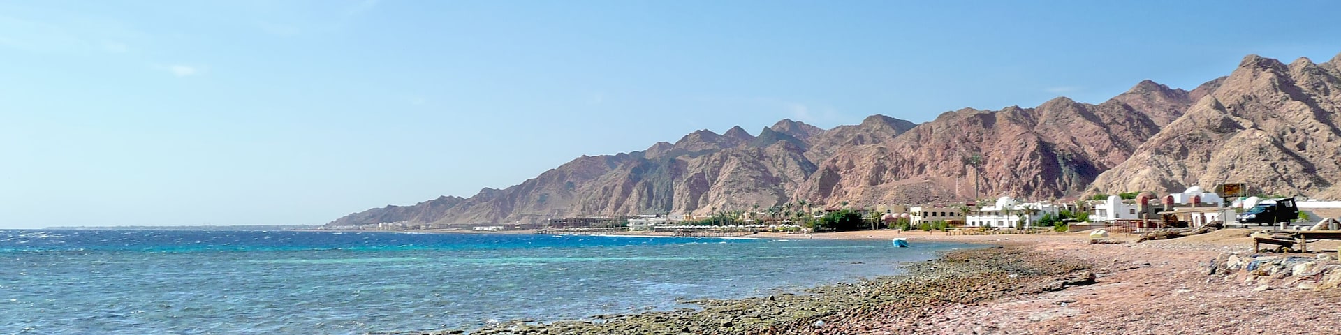 Coastal landscape north of Dahab, Sinai, Egypt, looking south — sunlit shoreline with distant buildings and the rugged mountain range rising beyond the Red Sea