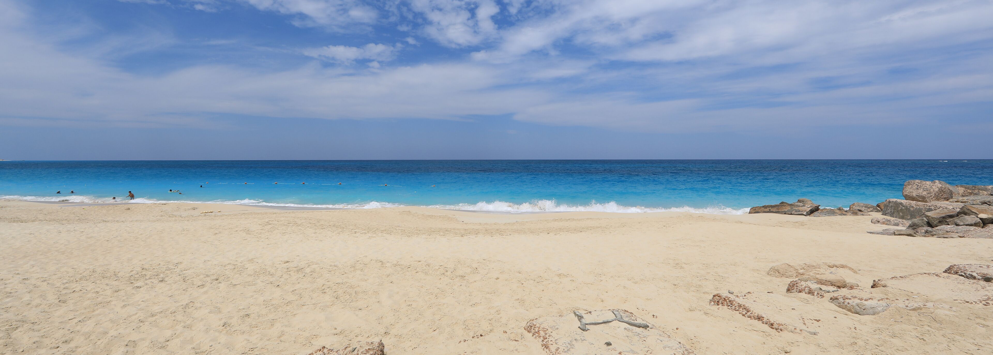A sandy beach with clear blue water and scattered rocks.