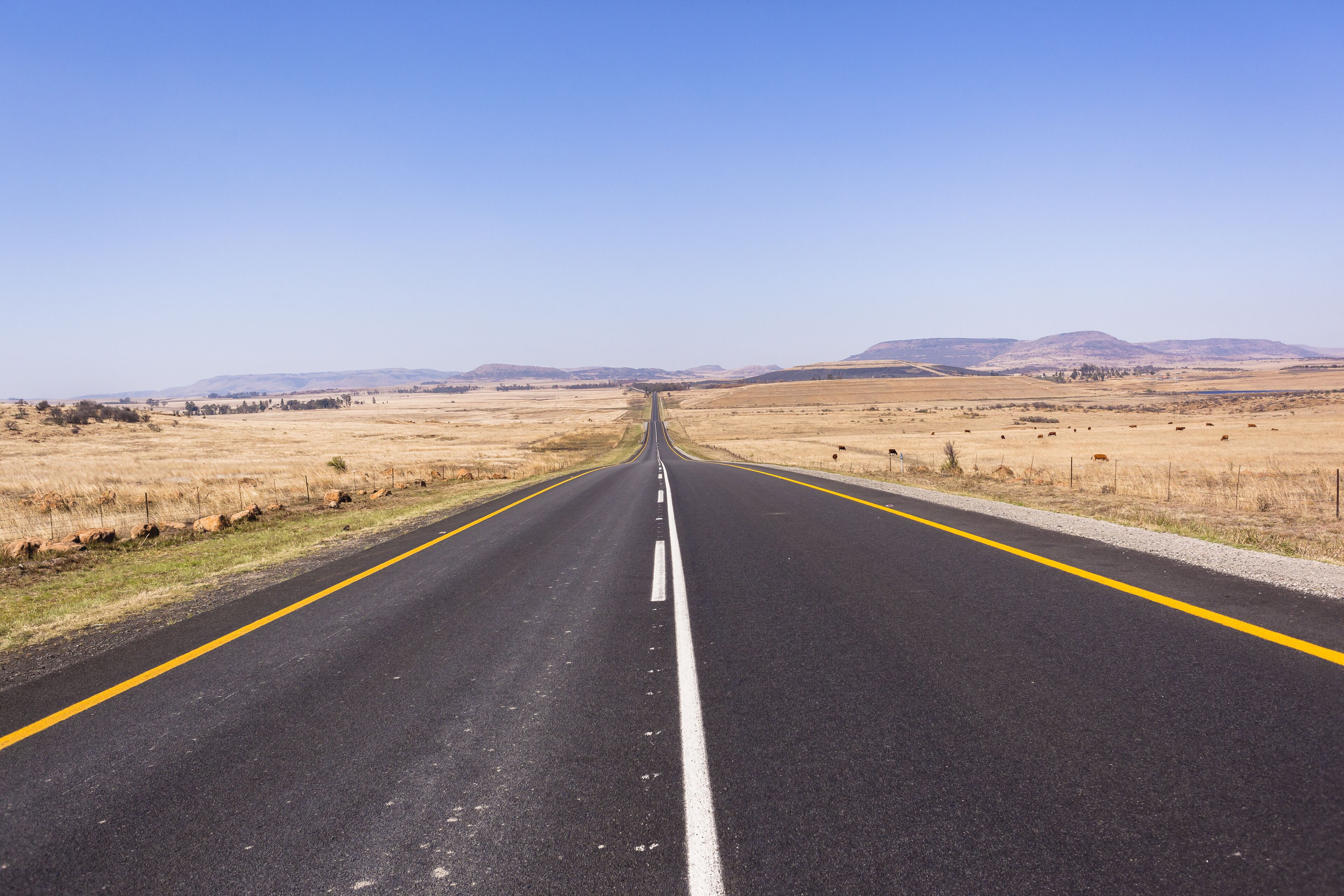 A straight road with yellow lines, flanked by dry grasslands and distant mountains under a clear sky.