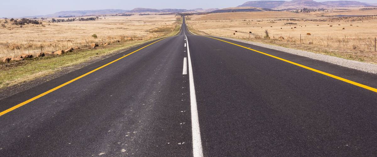 A straight road with yellow lines, flanked by dry grasslands and distant mountains under a clear sky.