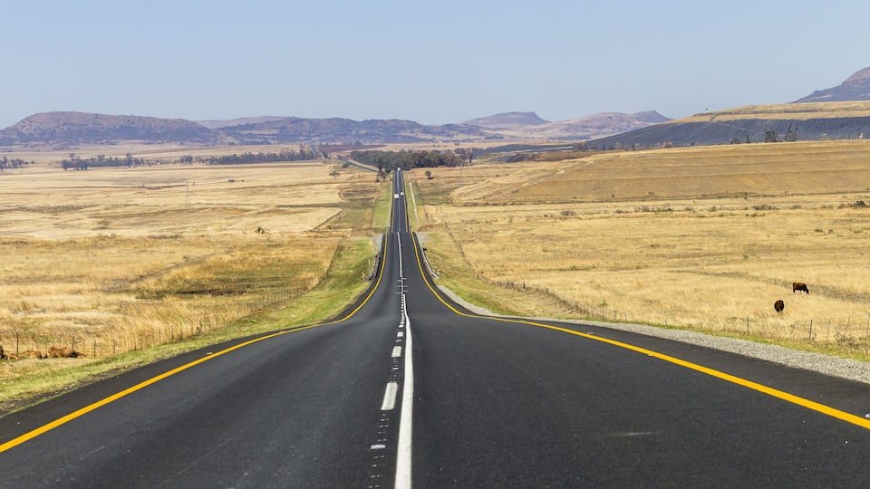 A straight road with yellow lines, flanked by dry grasslands and distant hills.