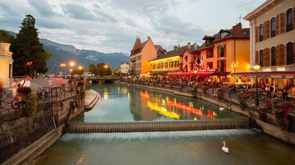 A riverside town with a swan swimming, buildings with restaurants, and mountains in the background.