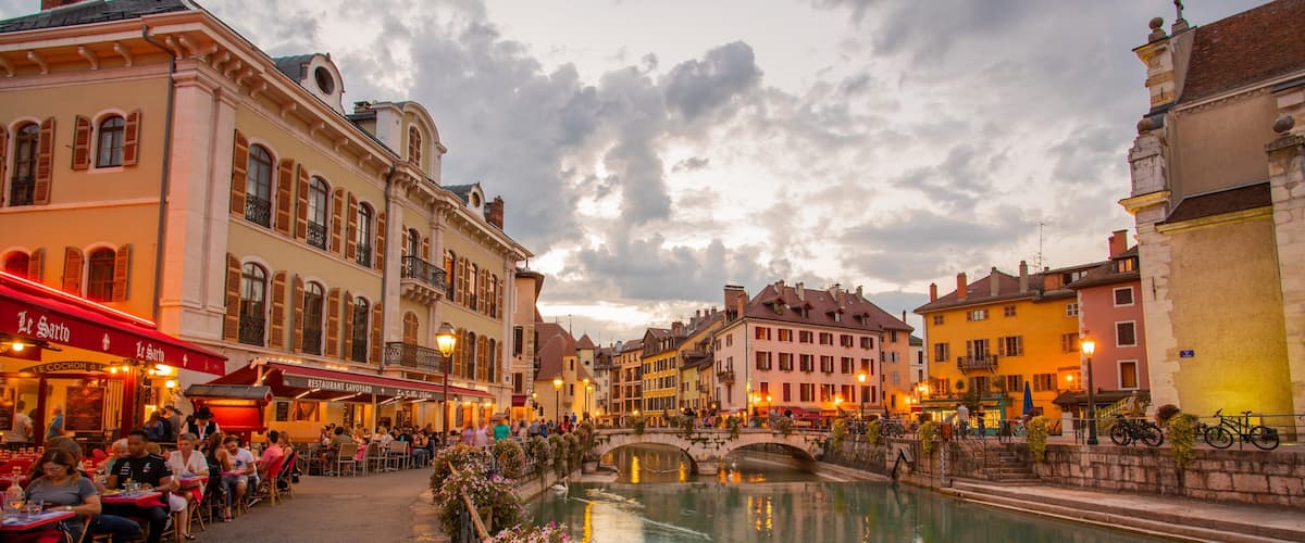 A riverside café area with outdoor seating, historic buildings, and a stone bridge.