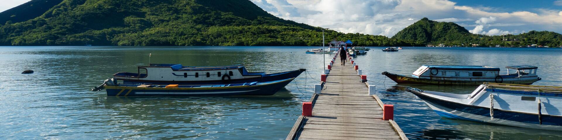 A wooden pier leading to boats on calm water with a green mountain in the background.