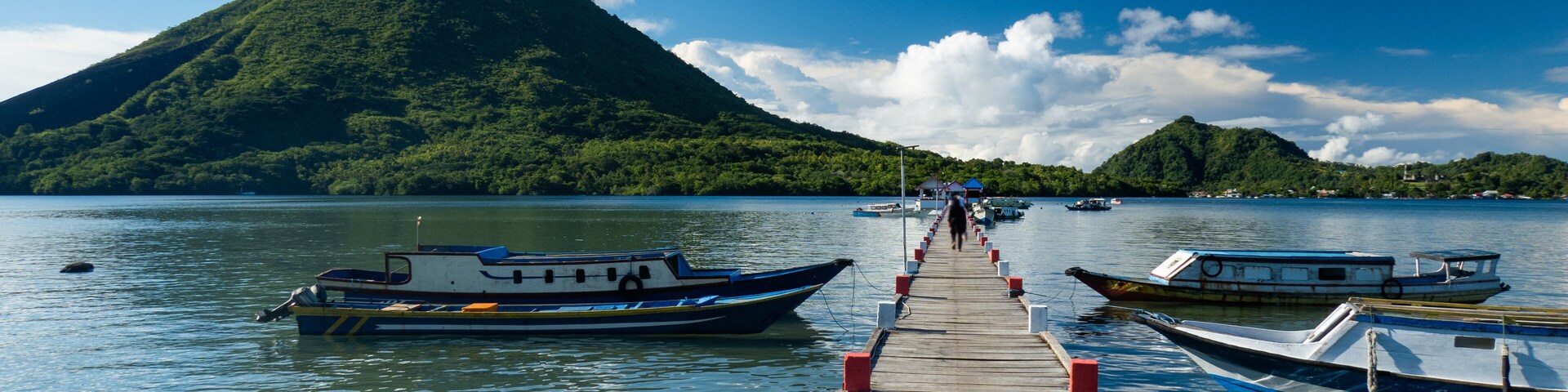 Un muelle de madera que lleva a botes sobre aguas tranquilas, con una montaña verde al fondo.