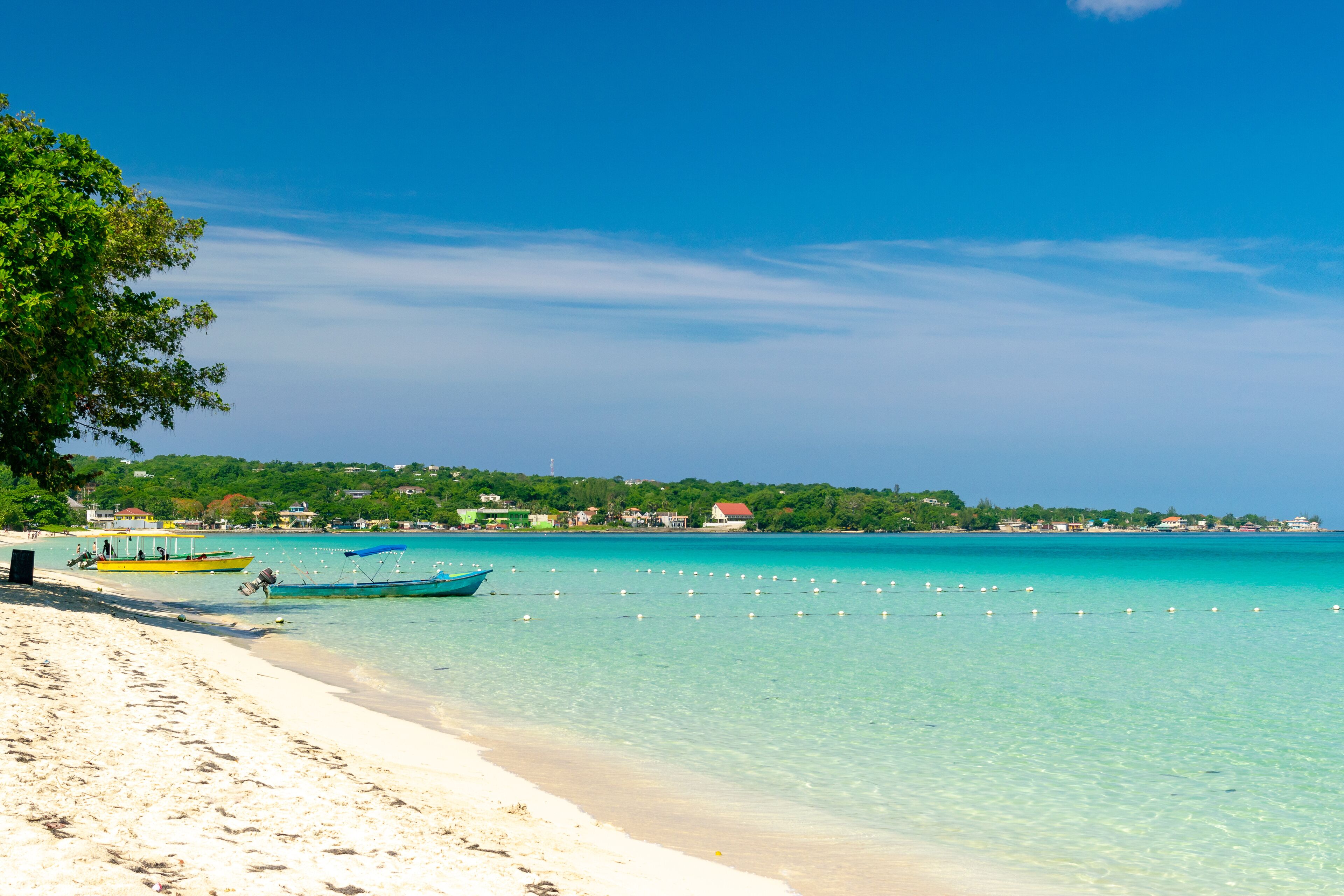A beach with clear turquoise water, boats, and a distant shoreline with buildings.