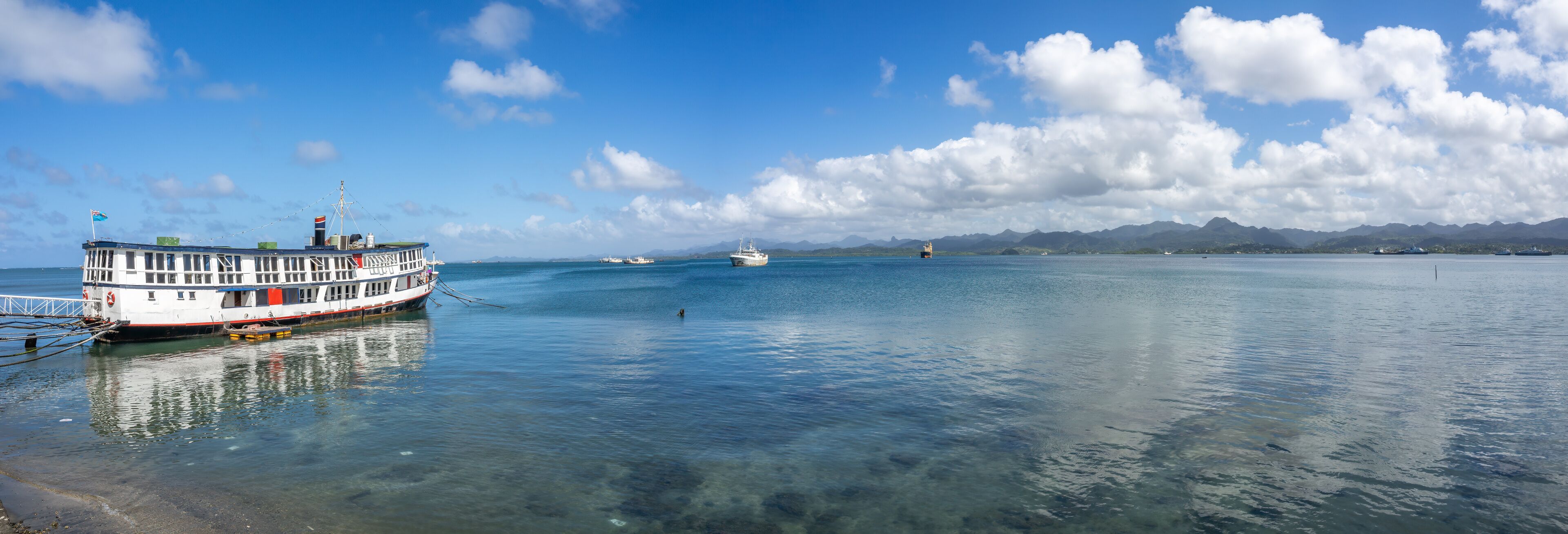 A large ferry docked at a harbor with clear blue water and distant mountains under a partly cloudy sky.