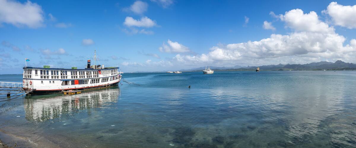 A large ferry docked at a harbor with clear blue water and distant mountains under a partly cloudy sky.