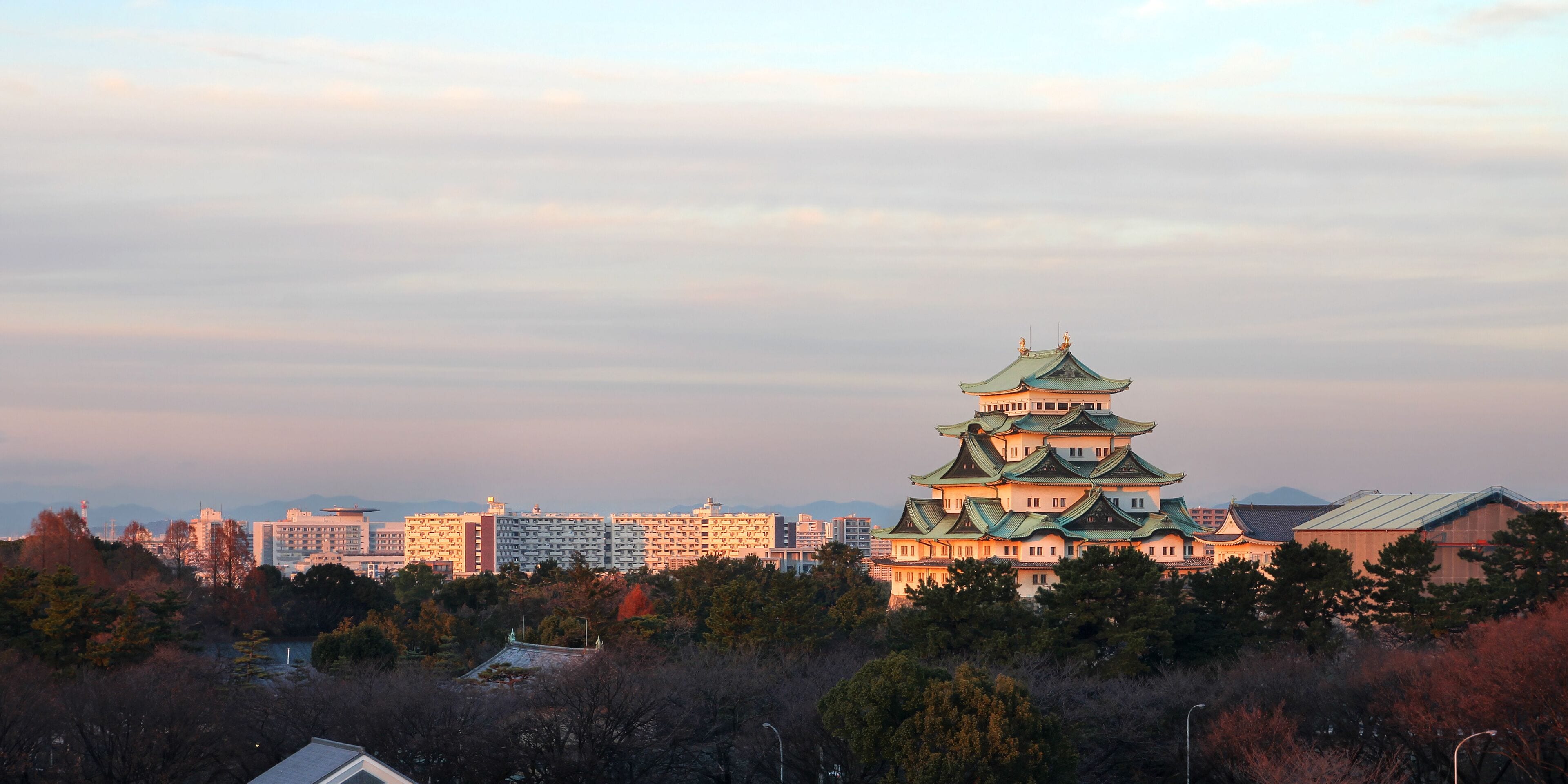 A traditional Japanese castle with multiple tiers and green roofs, surrounded by modern buildings and trees.