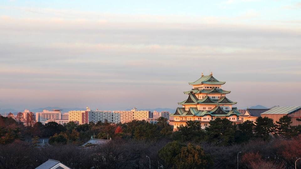 A traditional Japanese castle with multiple tiers and green roofs, surrounded by modern buildings and trees.