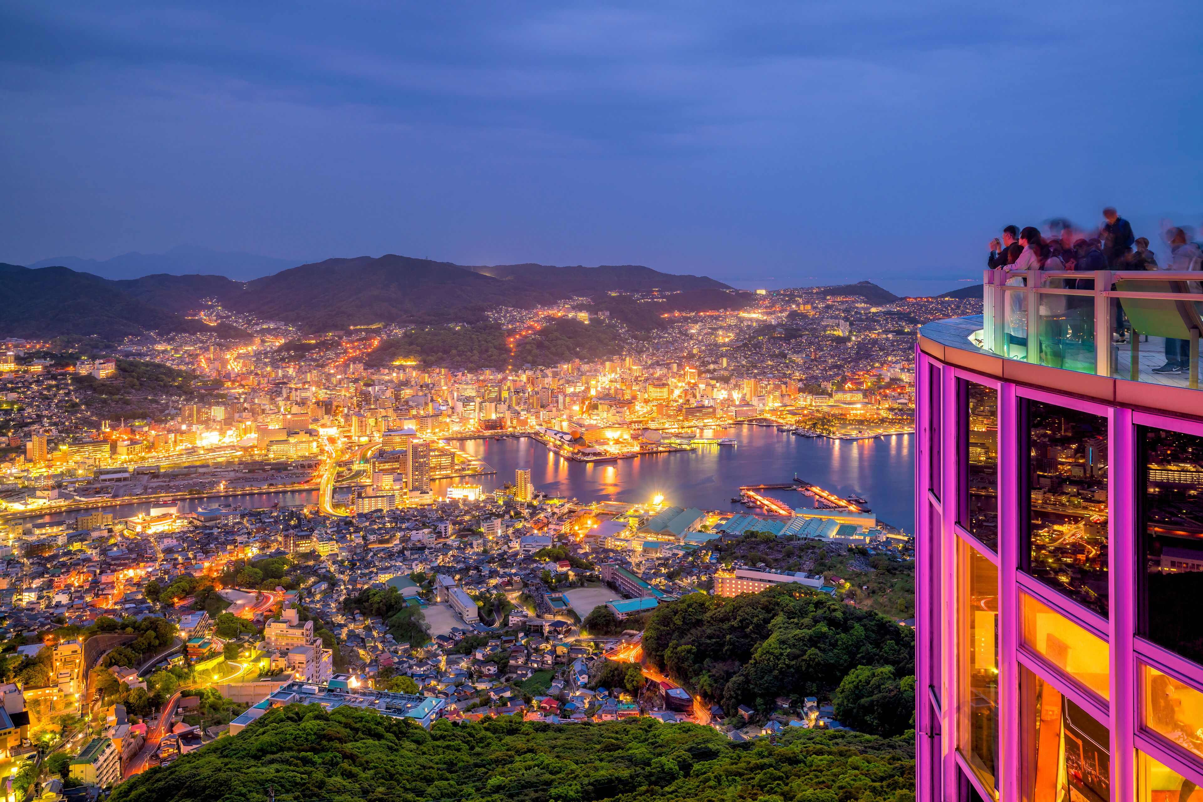 A panoramic cityscape at dusk with a river, bridges, and a high-rise building with a glass observation deck.