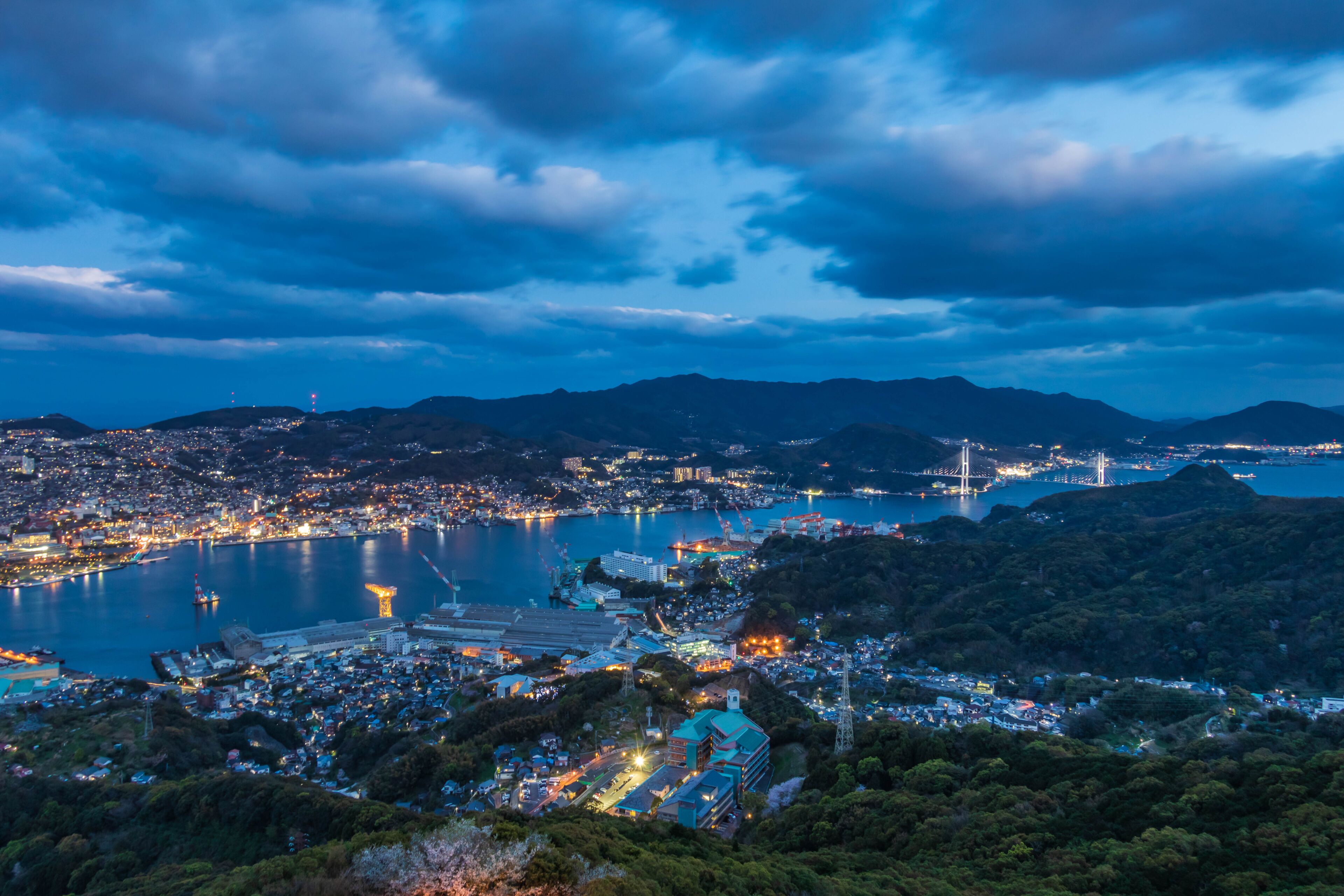 A coastal cityscape at dusk with a prominent bridge, a bay, and surrounding hills.