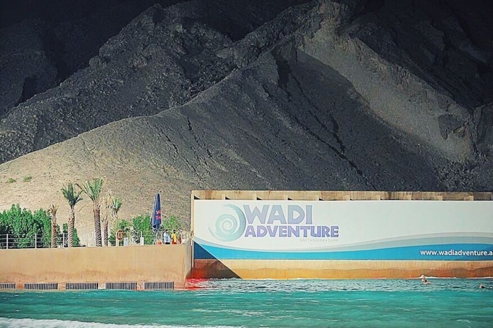 A beach with clear turquoise water, a large sign for WADI ADVENTURE, and a mountainous backdrop.