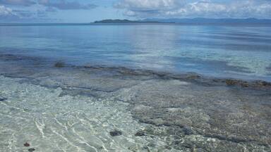A clear tropical beach with shallow waters, visible rocks, and a distant island.