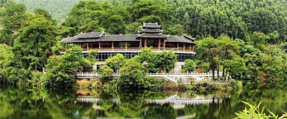 A traditional wooden building with a tiled roof, surrounded by lush greenery and a calm lake.