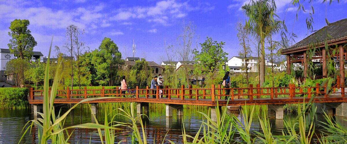A wooden bridge over a calm water body, surrounded by lush greenery and trees.