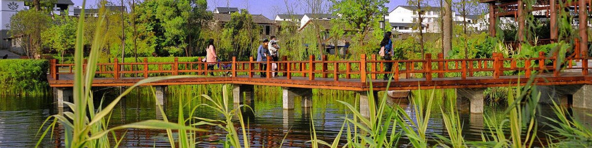 A wooden bridge over a calm water body, surrounded by lush greenery and trees.
