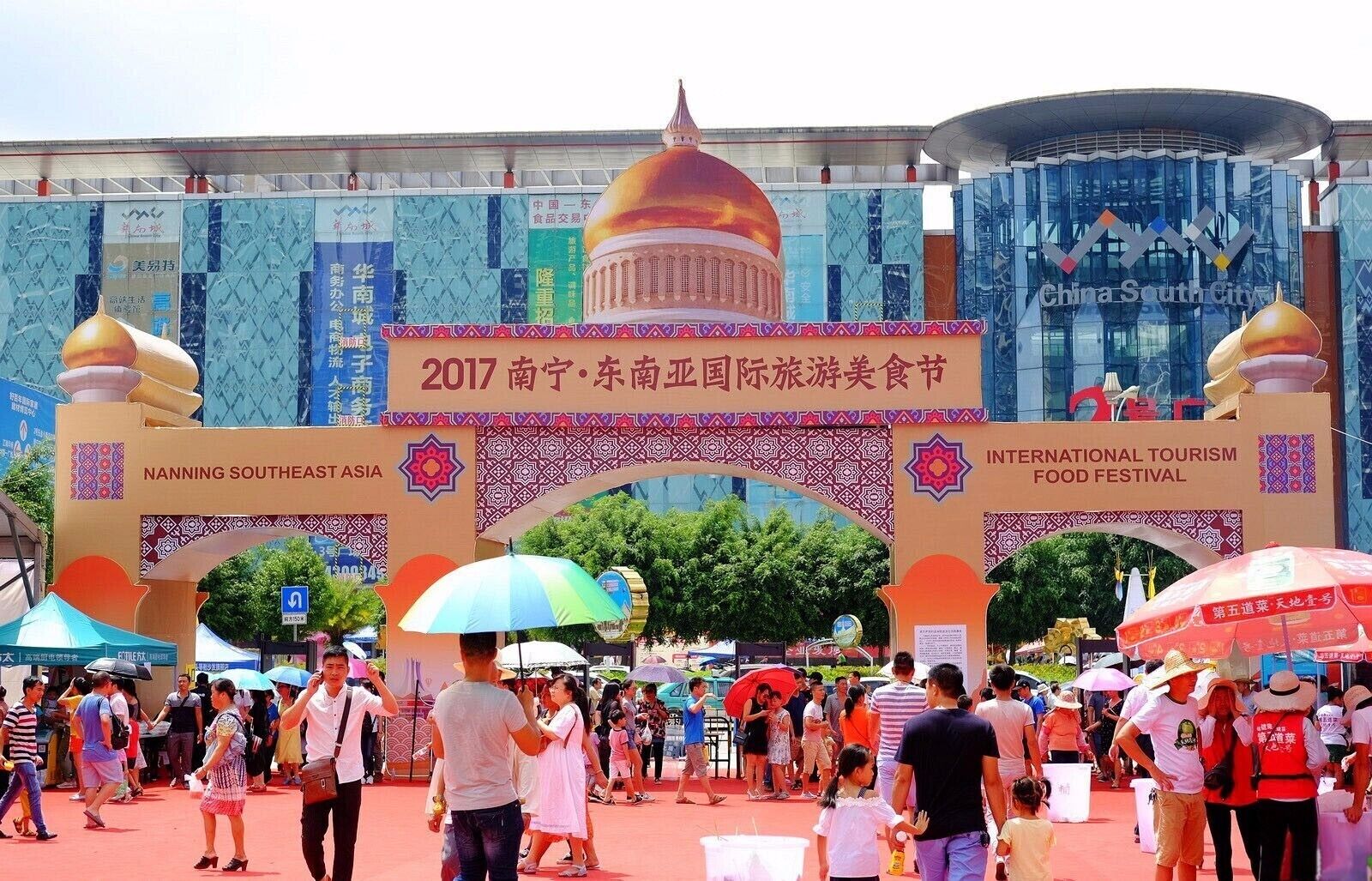 Una entrada concurrida al Festival Internacional de Turismo y Gastronomía del Sudeste Asiático en Nanning, con un arco grande y sombrillas de colores.
